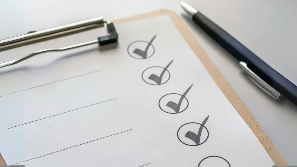 A close-up shot of a light-colored clipboard with a document that has four checkboxes marked with dark checkmarks. To the right of the clipboard, a dark pen rests on the surface, angled slightly. The background is a plain, light surface. The document on the clipboard also has faint horizontal lines, suggesting a form or list.